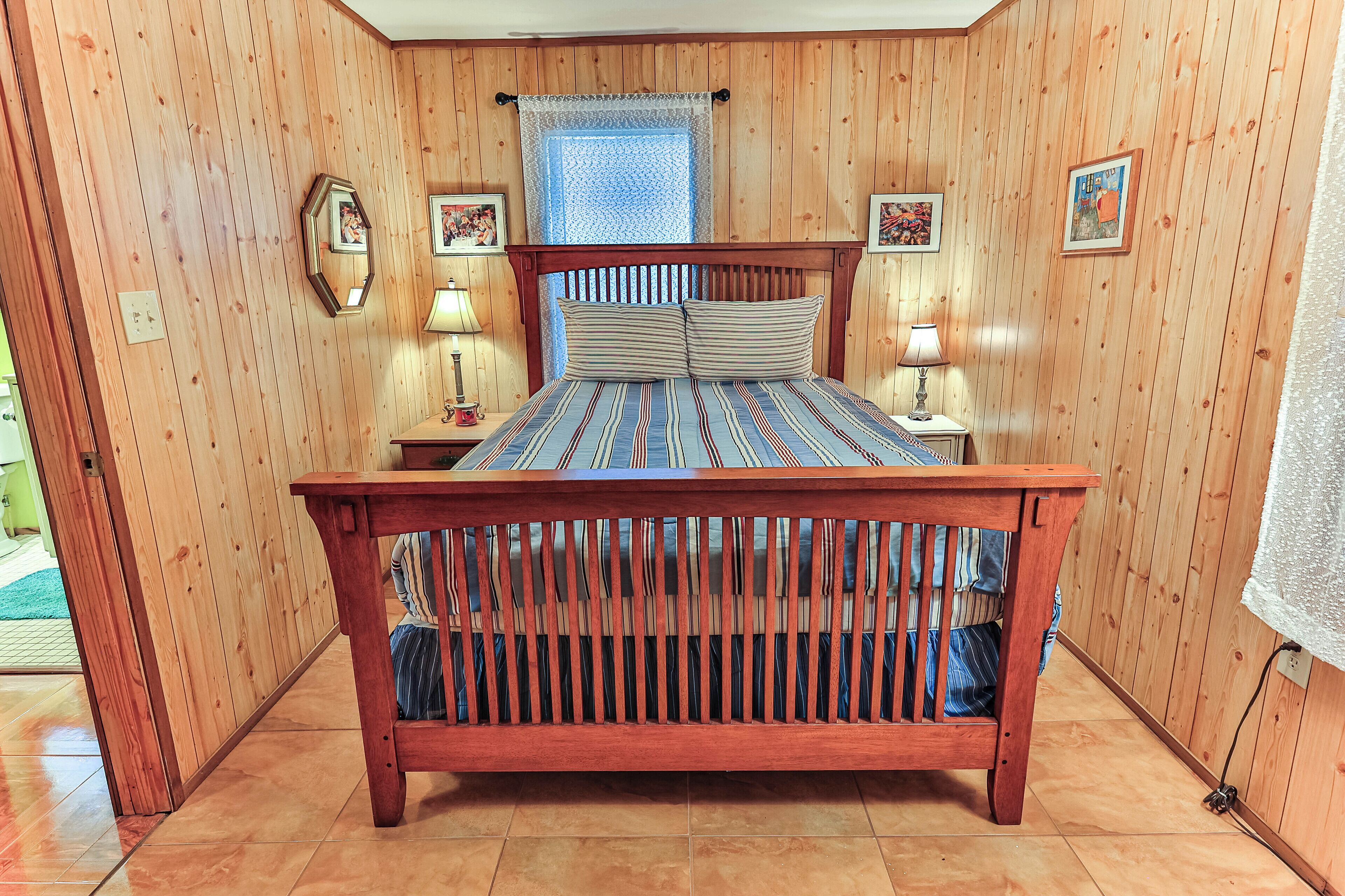 A wooden-paneled room with a bed, two bedside lamps, a window with a lace curtain, and framed pictures on the walls.