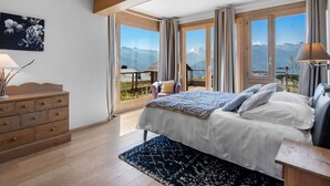 Un dormitorio con una cama grande, cómoda de madera y vistas a las montañas a través de las ventanas.