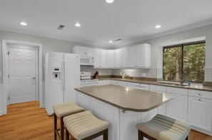 Una cocina moderna con armarios blancos, una isla grande con taburetes y un ventanal con vista a árboles.