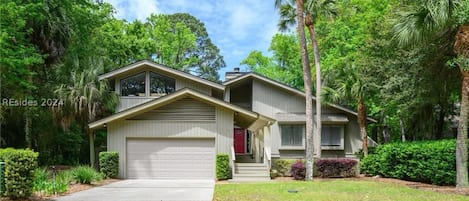 A two-story house with a garage, surrounded by lush greenery and palm trees.