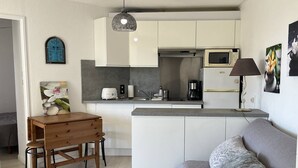 A compact kitchen with white cabinets, a microwave, and a coffee maker. There's a wooden table with two stools and a grey sofa with a textured throw blanket.