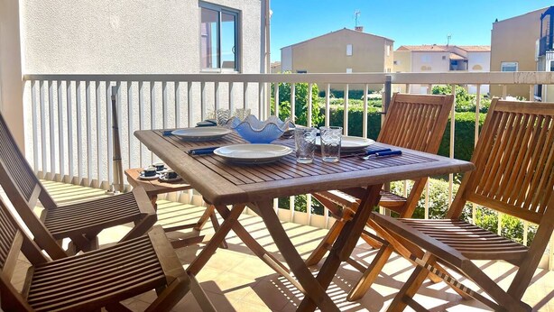 A balcony with a wooden table set for dining, two wooden chairs, and a view of neighboring buildings.