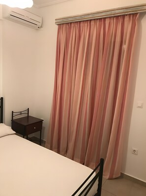 Een slaapkamer met een bed, nachtkastje, stoel, raam met roze gordijnen en een airconditioning.