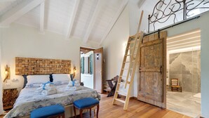 A bedroom with a wooden headboard, a bed with blue bedding, two matching chairs, a wooden ladder, and a large wooden door leading to a bathroom.