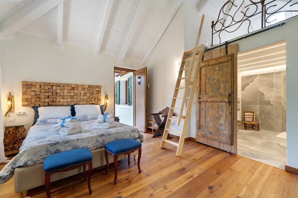 Un dormitorio con cabecera de madera, una cama con ropa de cama azul, dos sillones a juego, una escalera de madera y una amplia puerta de madera que da a un baño.