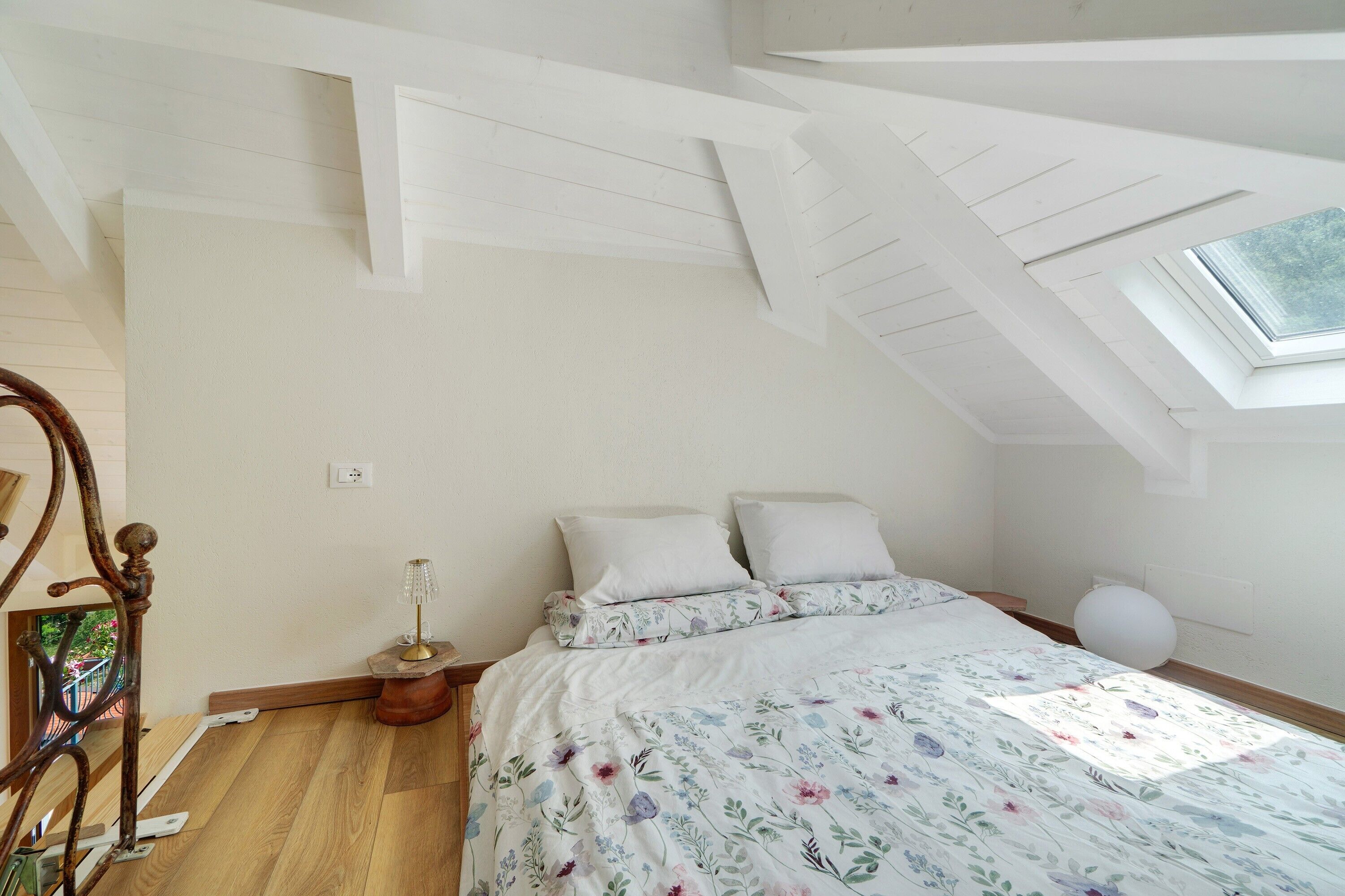 Une chambre avec une lucarne, un lit recouvert d’une couette à motifs floraux, une table de chevet avec une lampe et un plancher en bois.