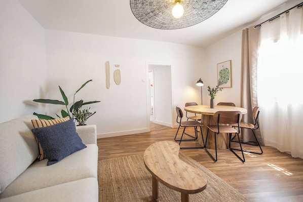 A living room with a sofa, a coffee table, a dining table with chairs, a potted plant, and a ceiling light fixture.
