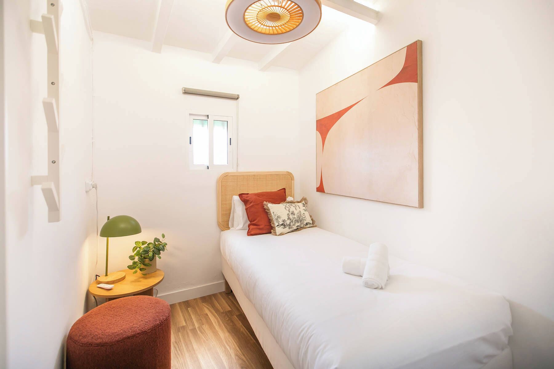 A small, well-lit bedroom with a single bed, a nightstand with a lamp, a potted plant, and a large abstract painting on the wall.