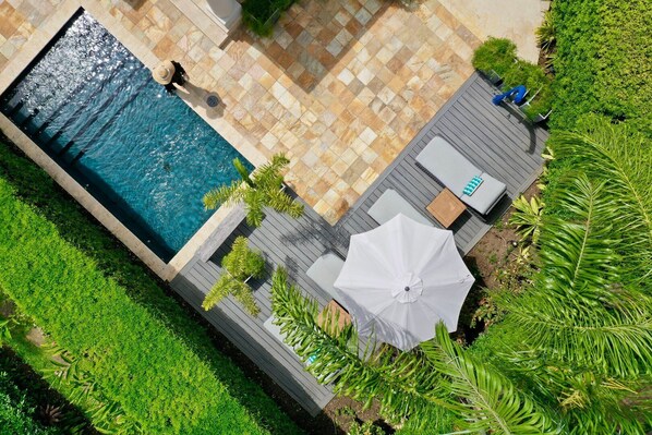 An aerial view of a swimming pool with tiled flooring, surrounded by lush greenery and a wooden deck area with seating and an umbrella.