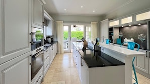 A modern kitchen with white cabinetry, a black countertop, and a central island. There are blue chairs and a glass-top table. - Tropical Holiday Rental Villa - Villatre (Mullins)