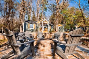 A cabin with a stone fire pit, surrounded by Adirondack chairs, set in a wooded area.