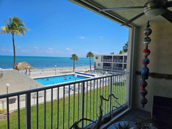 Un balcó amb vista a una piscina i una platja, una cabana de palla i un edifici amb balcons.