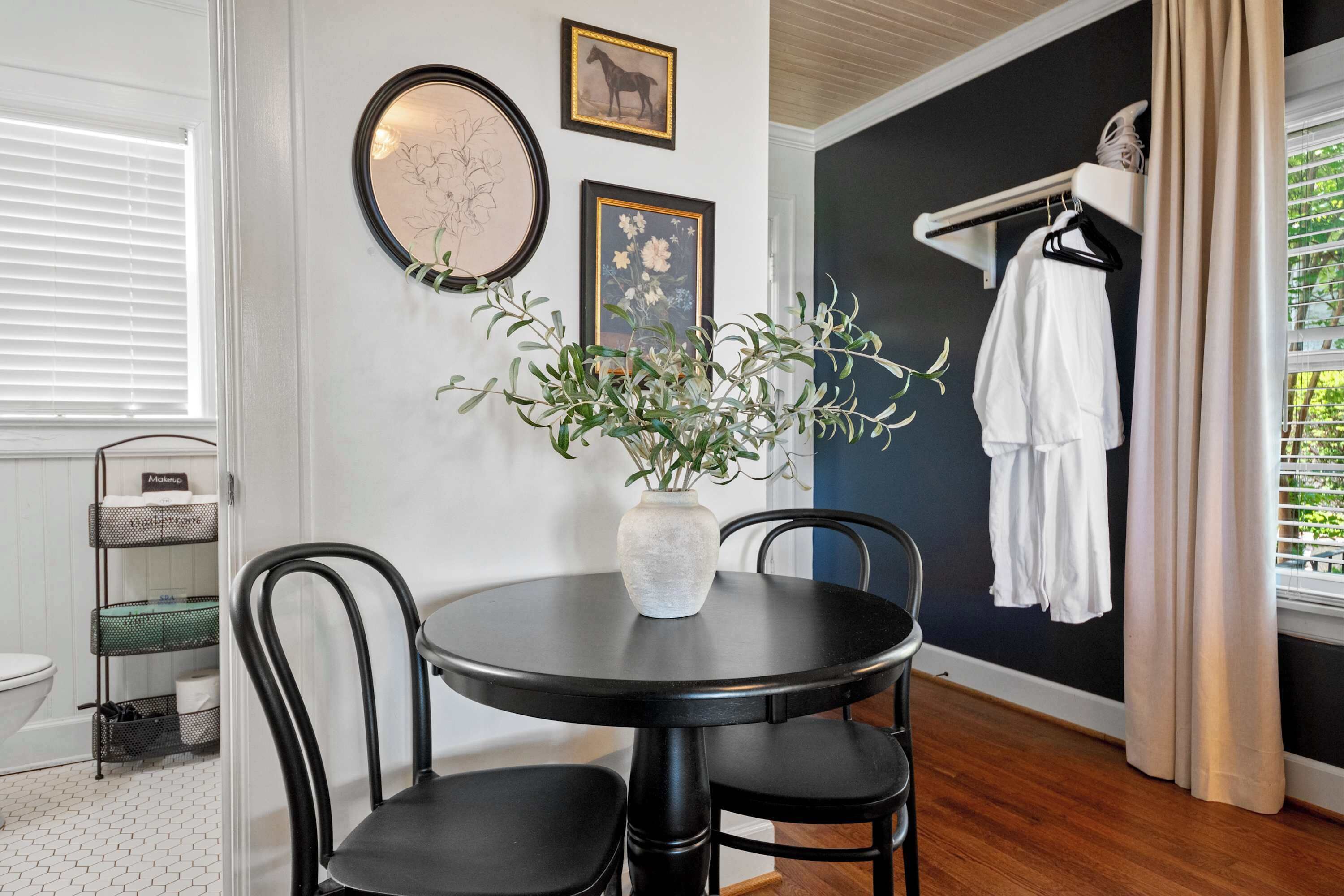 A room with a round table, chairs, a vase with plants, a rack with clothes, and a window with curtains.