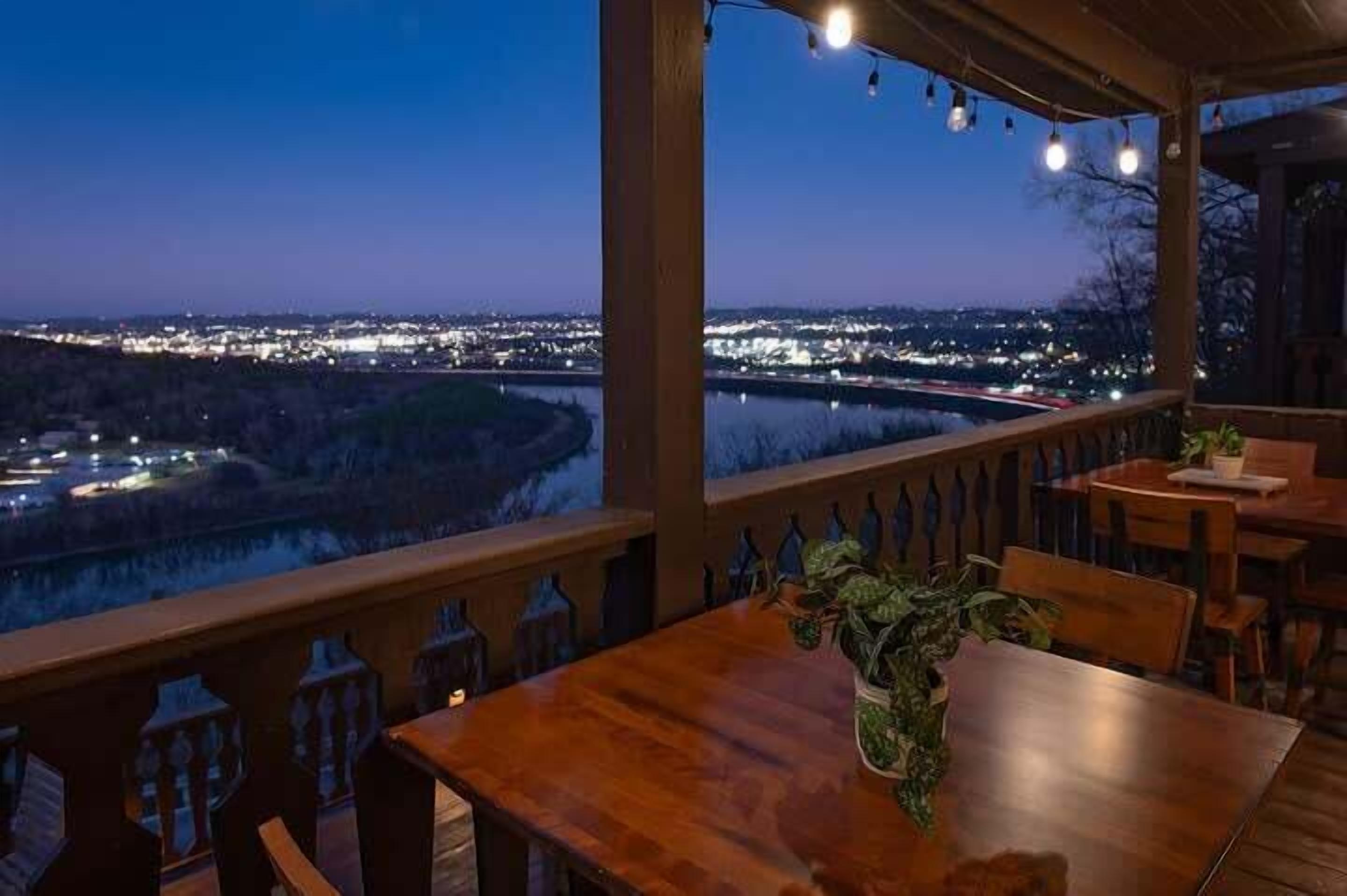 A wooden deck with a table and chairs, overlooking a river and city lights at dusk.