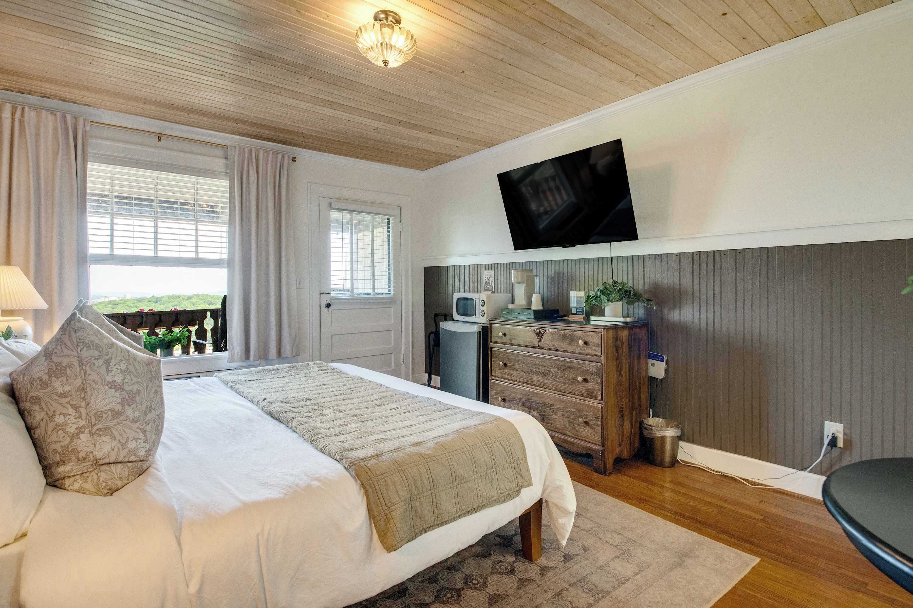 A bedroom with a bed, a television mounted on the wall, a dresser, and a window with curtains.