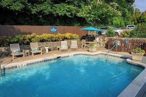 A swimming pool with lounge chairs and umbrellas, surrounded by a wooden fence and greenery.
