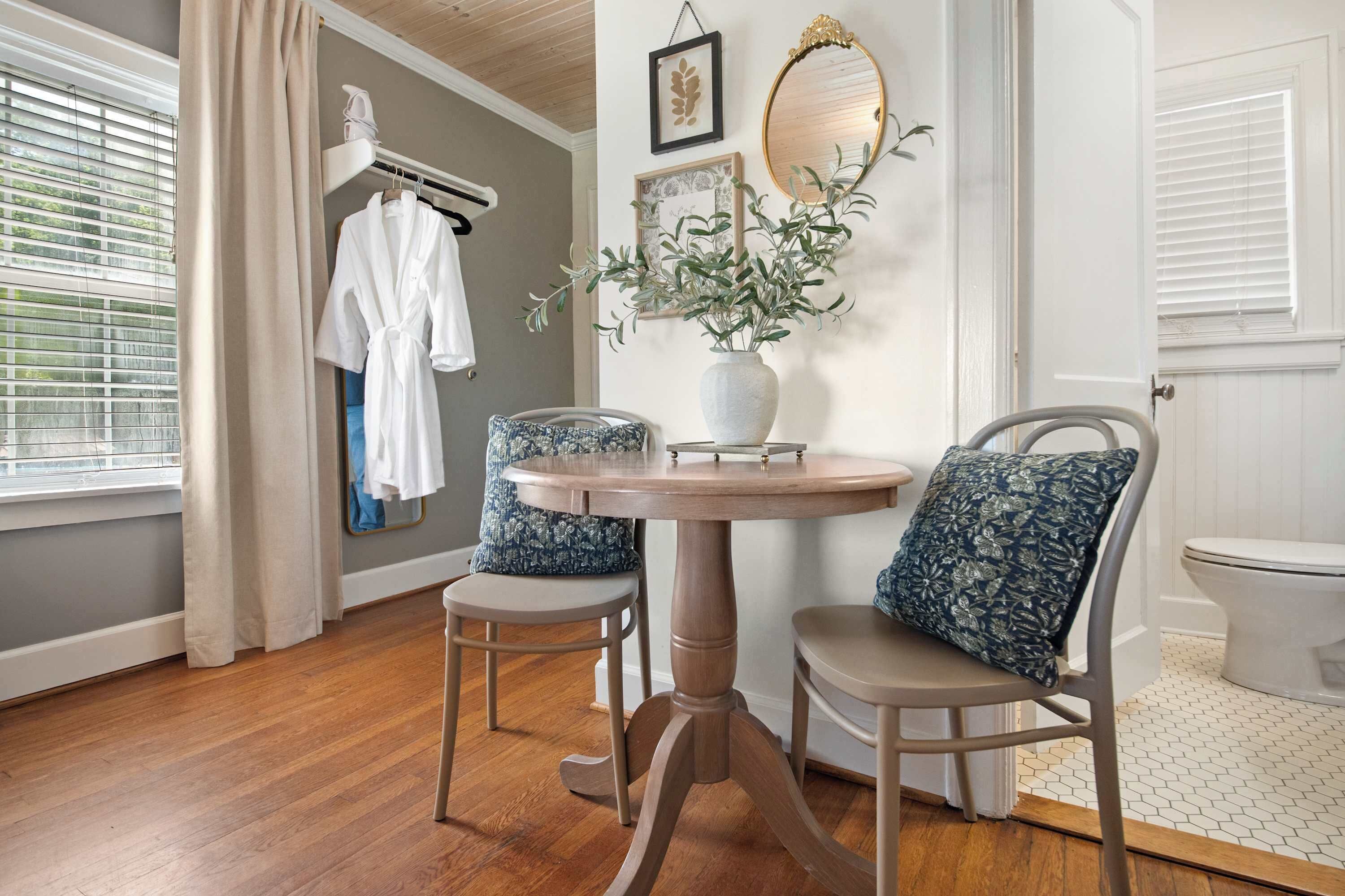 A room with a round wooden table, two chairs with floral cushions, a white robe on a rack, a vase with greenery, a mirror, and a toilet.