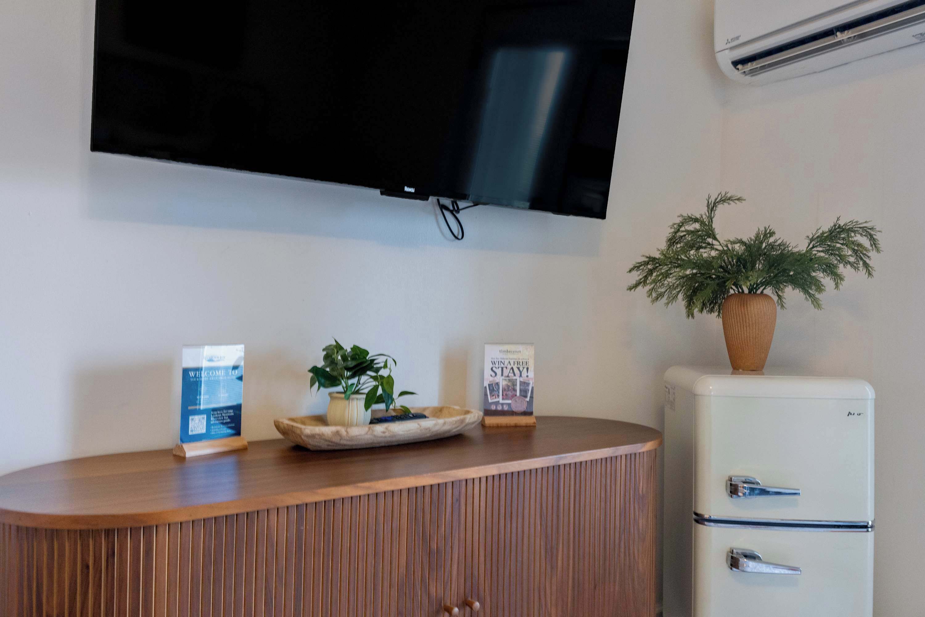 Un mostrador de recepción con un televisor de pantalla plana, una planta en maceta, un cartel de bienvenida y un refrigerador de estilo vintage.