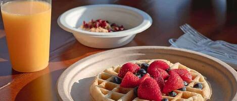 A breakfast setting with waffles topped with berries, a glass of orange juice, and a bowl of cereal on a wooden table with a scenic view.