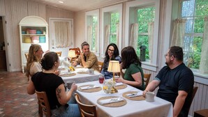 Un coin repas avec des tables dressées pour le repas, un buffet blanc contenant de la vaisselle, et une vue sur la verdure à travers les fenêtres.