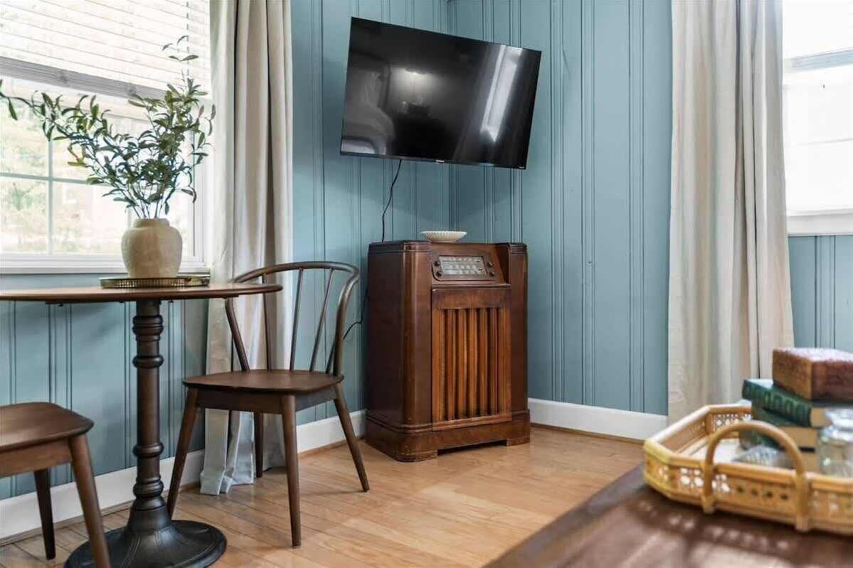 A room with a wooden table, a chair, a vintage radio, and a television mounted on the wall.