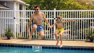 Dos personas saltando a una piscina con una cerca blanca y una casa al fondo.