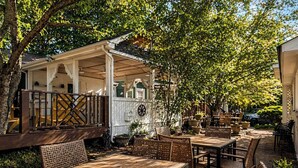Un patio al aire libre con mesas de madera y sillas de mimbre, rodeado de follaje y un edificio blanco con porche.