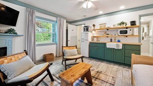 Una habitación acogedora con cama, un banco de madera, una silla, un televisor, un ventilador de techo, una ventana con cortinas y una kitchenette con armarios verdes y un microondas.