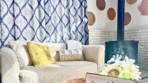 Una sala de estar acogedora con un sofá estampado, cojines decorativos, una mesa de centro de madera con un cartel que dice "wifi happy getaway" y una pared de ladrillo rústica.