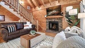 A cozy log cabin interior with a wooden staircase, a fireplace, a brown leather sofa, and a large window overlooking a forest. - Smoky Mountain Summer Fun Near Dolly! Pool + Arcade + Gaming Wall! (Sevierville)
