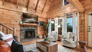 A cozy log cabin interior with a fireplace, comfortable seating, and a large window with curtains. - Smoky Mountain Summer Fun Near Dolly! Pool + Arcade + Gaming Wall! (Sevierville)