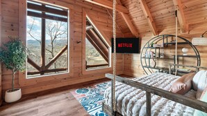 A cozy wooden cabin room with a bed, a bookshelf, and a large window offering a view of the outdoors. - Smoky Mountain Summer Fun Near Dolly! Pool + Arcade + Gaming Wall! (Sevierville)