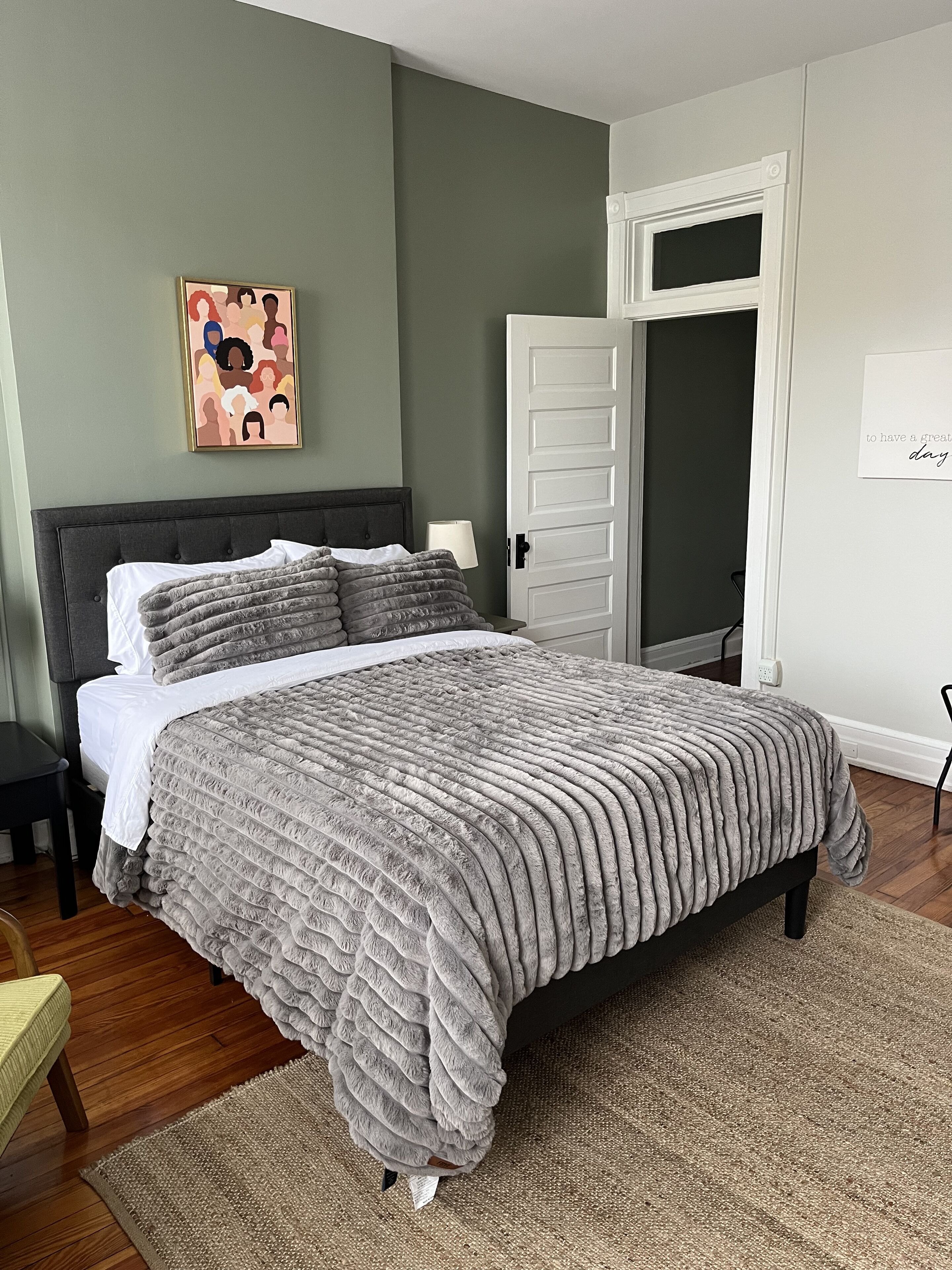 A bedroom with a large bed, a nightstand, a chair, a door, and a wall with a framed picture.
