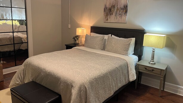 A neatly made bed with a headboard, a bedside table with a lamp, a small bench, and a view through a window with a frosted glass panel.