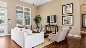 Ein Wohnzimmer mit einer Couch, Sesseln, einem Couchtisch und einem Fernseher. Es gibt ein Fenster mit Jalousien, eine Pflanze und gerahmte Kunstwerke an der Wand.
