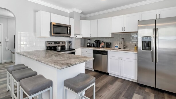 Una cocina moderna con armarios blancos, electrodomésticos de acero inoxidable, encimera de mármol y una zona con taburetes de bar.