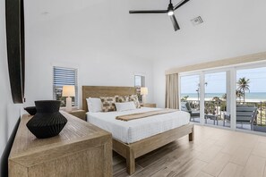 A modern bedroom with a large bed, a wooden bedside table, a black vase, and a view of the beach through a sliding glass door. - Prime Location, Heated Pool, Game Room - FMB- Beach Please - Roelens (Fort Myers Beach)