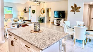 A modern kitchen with granite countertops, white cabinetry, and a central island. There is a dining area with white chairs and a glass table. A television is mounted on the wall, and there are ceiling fans above. - Tropical Oasis, Heated Pool & Spa, Bikes - Villa 7 Palms - Roelens (Cape Coral)