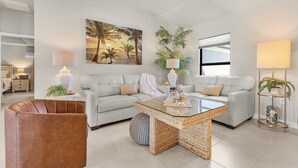 A modern living room with a light-colored sofa, a glass coffee table, and a brown leather chair. There is a large painting of palm trees on the wall, a potted plant, and a floor lamp. - Tropical Oasis, Heated Pool & Spa, Bikes - Villa 7 Palms - Roelens (Cape Coral)