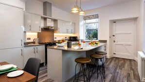 A modern kitchen with white cabinetry, a central island with stools, and a dining area with a wooden table and chairs. - The Firs - Centre of Bowness, parking for 5+ cars (Bowness-on-Windermere)