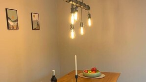 A dining area with a wooden table, two framed artworks, a hanging light fixture, a candle, a fruit bowl, a loaf of bread, and a bottle of wine.