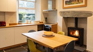 A kitchen with a stone fireplace, a dining table with chairs, and a window with a view of greenery. - Holmdale - Central Ambleside (Ambleside)