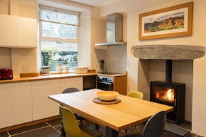A kitchen with a stone fireplace, a dining table with chairs, and a window with a view of greenery.