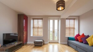 Ein modernes Wohnzimmer mit einem Flachbildfernseher, einem grauen Sofa mit bunten Kissen, Holzboden und einer roten Akzentwand.