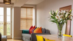 Ein modernes Wohnzimmer mit einem grauen Sofa, bunten Kissen, einem Holzesstisch mit gelben Stühlen und einer Vase mit weißen Blumen.