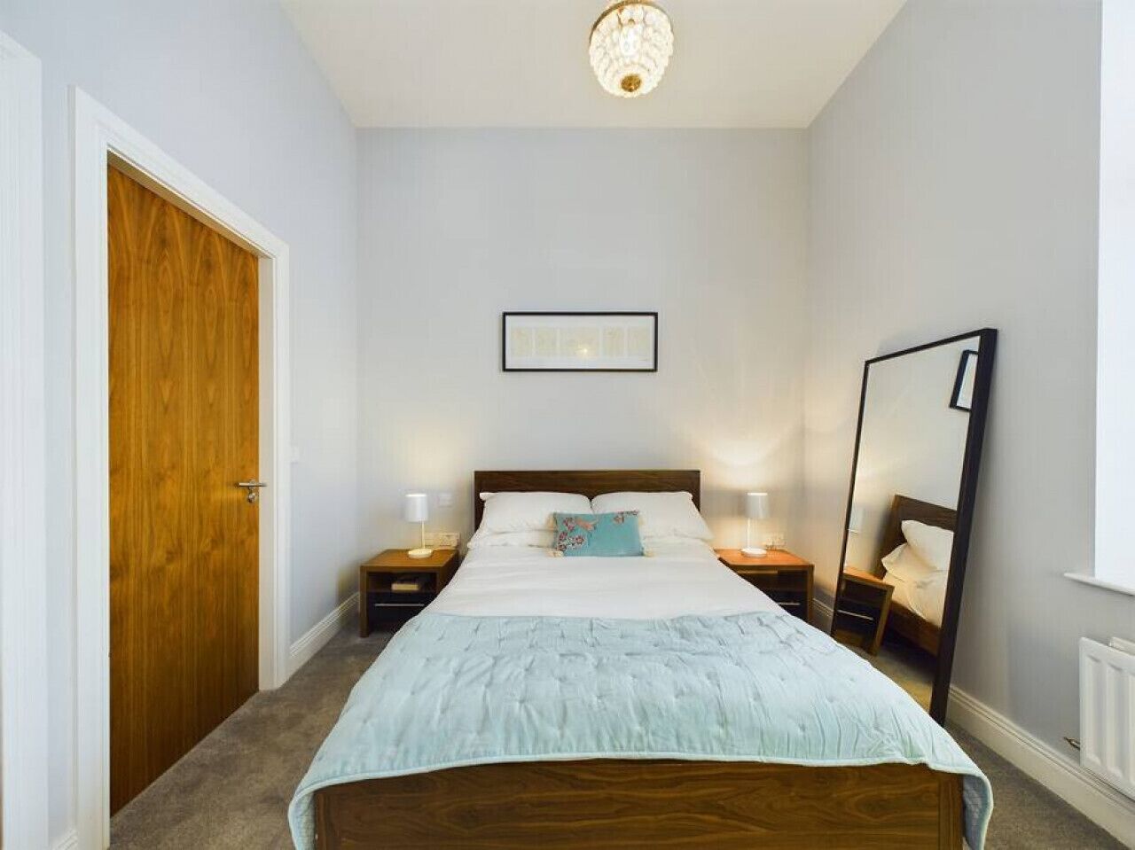 A bedroom with a bed, two bedside tables, a nightstand, a wooden door, a framed picture on the wall, and a mirror on the wall.
