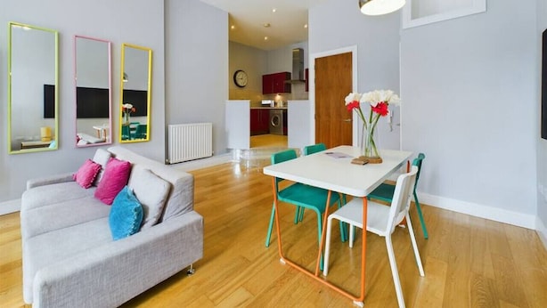 A modern living room with a grey sofa, colorful cushions, a dining table with chairs, and a kitchen in the background.