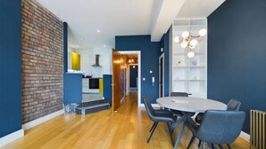 Uma cozinha moderna com parede de destaque em tijolo, área de jantar com mesa e cadeiras redondas, e uma estante branca em frente a uma parede azul escura.