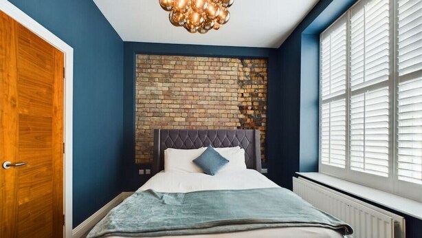 Quarto com parede de tijolos, cabeceira estofada, cama com roupa de cama branca, manta azul e luminária suspensa.