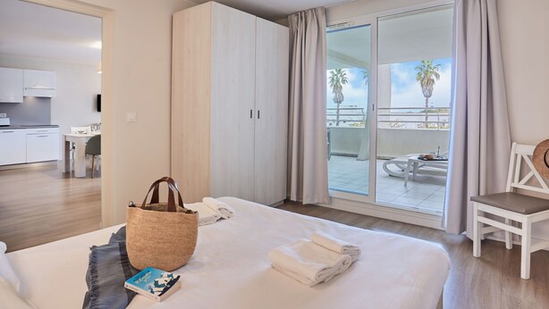 A hotel room with a bed, a bedside table, a chair, a balcony with a view, and a kitchenette. - Odalys Référence Les Dunes du Soleil by Interhome (La Grande Motte)